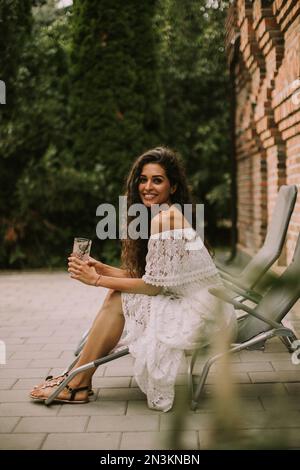 Bella giovane donna con capelli ricci si rilassa su una sedia a sdraio e gode di un bicchiere di limonata fresca Foto Stock