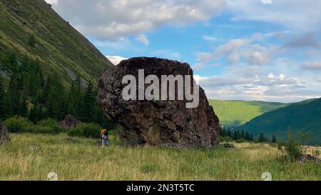 Una ragazza viaggiatore con uno zaino in occhiali e un cappello cammina appoggiato su pali da trekking, sullo sfondo di una grande pietra nelle montagne Altai o Foto Stock