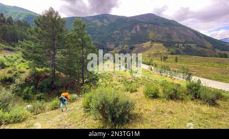 Ragazza viaggiatore con uno zaino su bastoni da trekking che sale sul ripido sentiero di pietra di Altai sullo sfondo delle case. Foto Stock