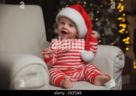 Carino bambino in cappello di Santa e pigiama di Natale brillante che tiene canna caramella a casa Foto Stock