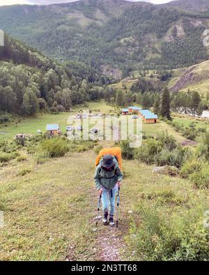 Ragazza viaggiatore con uno zaino su bastoni da trekking che sale sul ripido sentiero di pietra di Altai sullo sfondo delle case base. Foto Stock