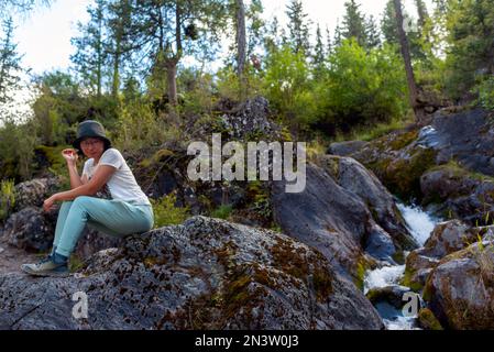 Una ragazza asiatica in un cappello e occhiali siede sorridendo su una pietra contro un torrente di montagna nella foresta. Foto Stock