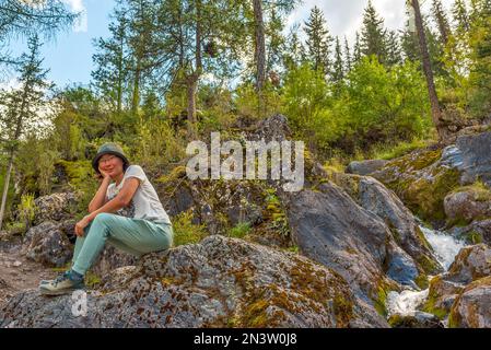 Una ragazza asiatica in un cappello e occhiali siede su una pietra sullo sfondo di un torrente di montagna nella foresta. Foto Stock