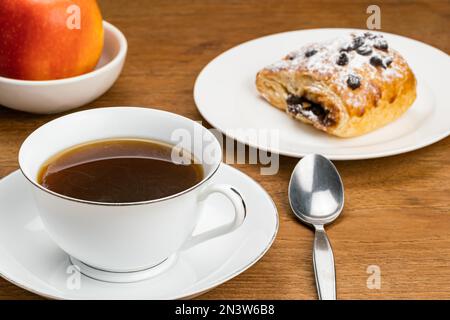 Caffè nero caldo in tazza di ceramica bianca su piattino di ceramica per colazione con pasticceria danese ripiena di crema di cioccolato con gocce di cioccolato e. Foto Stock