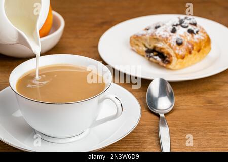 Versare il latte fresco nel caffè in una tazza di ceramica bianca su un piattino di ceramica con pasticceria danese ripiena di crema di cioccolato con gocce di cioccolato e. Foto Stock