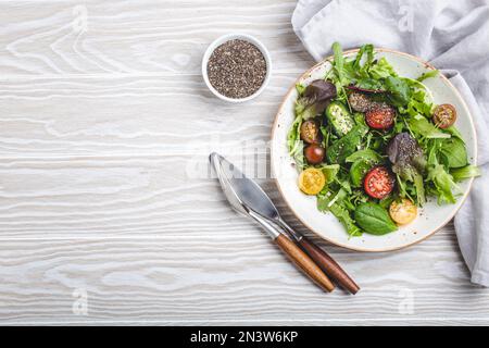 Verdure insalata sana vegetariana con pomodori ciliegini rossi gialli, foglie verdi e semi di chia su piatto bianco su fondo rustico in legno bianco Foto Stock