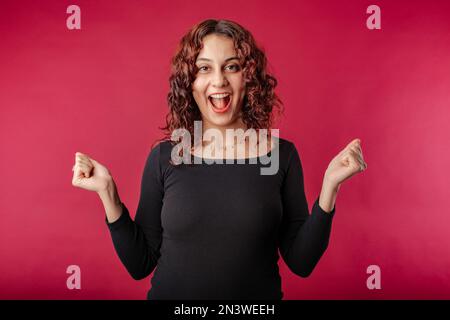 Giovane ragazza rossa sorridente felice isolato su mani di sfondo rosso in pugni grida di gioia e ride alla fotocamera. Vincere e festeggiare, trionfare Foto Stock