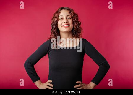 Bella donna rossa vestito nero in piedi isolato su sfondo rosso gode parlare e ride molto. Tocca il mento con la mano A. Foto Stock