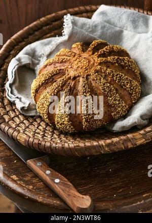 Pane rotondo di orzo e farina di mais appena sfornato con tovagliolo in vimini in un vecchio sgabello di legno d'annata. Cibo sano fatto in casa Foto Stock