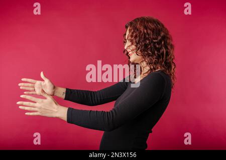 Vista laterale della giovane donna rossa in piedi isolata su sfondo rosso che stretching le mani come se dando per libero, offrendo di abbracciare, compiacenza e. Foto Stock