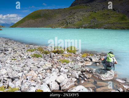 Una ragazza turistica con uno zaino si siede su una spiaggia di pietra vicino all'acqua del lago alpino turchese Karakabak nelle montagne Altai. Foto Stock