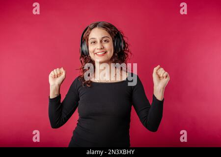 Donna millennial carina vestito nero a coste in piedi isolato su sfondo rosso ascoltare musica con cuffie, danza. Ballare con i musi preferiti Foto Stock