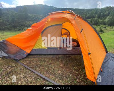 Una ragazza asiatica dopo il sonno si trova in una tenda arancione in un sacco a pelo sullo sfondo delle montagne. Foto Stock