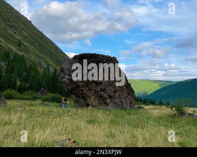 Una ragazza viaggiatrice con uno zaino in occhiali e un cappello cammina appoggiato su pali da trekking, sullo sfondo di una grande pietra nelle montagne Altai. Foto Stock