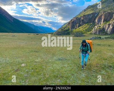 Una ragazza turistica da sola con uno zaino arancione e bastoncini da trekking cammina lungo una strada alpina in estate vicino alle montagne e al fiume Altai. Foto Stock