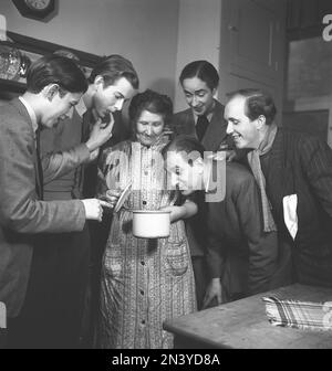 In cucina 1940s. Interno di una cucina e di una donna anziana che cucinano , con cinque uomini che vogliono ardentemente assaggiare il contenuto della pentola. Svezia 1945 Kristoffersson Ref M115-4 Foto Stock
