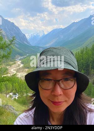 Una sorridente ragazza di viaggio asiatica in occhiali e un cappello porta un selfie al telefono sullo sfondo di un panorama di ghiacciai e montagne innevate in al Foto Stock