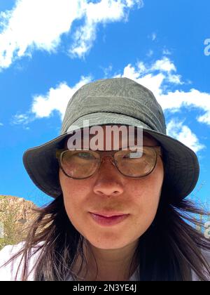 Una ragazza viaggiatore asiatica gioiosa in occhiali e un cappello prende un selfie al telefono sullo sfondo del cielo e delle rocce ad Altai in estate. Foto Stock