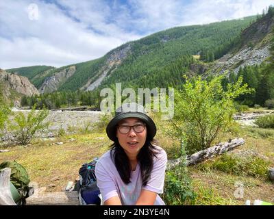 Una ragazza viaggiatore asiatica gioiosa in occhiali e un cappello prende un selfie al telefono sullo sfondo di un panorama delle montagne Altai in estate. Foto Stock