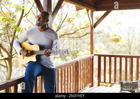 Un uomo anziano afro-americano sorridente che suona la chitarra mentre si siede sulla ringhiera nel balcone della cabina di tronchi Foto Stock