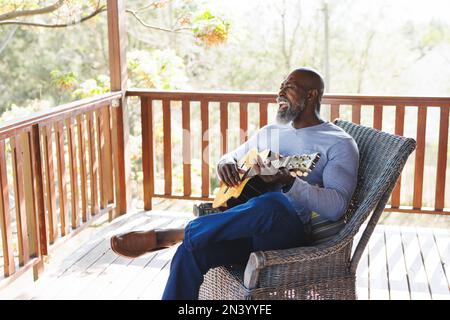 Calvo afroamericano anziano che suona la chitarra mentre si siede sulla sedia nel balcone alla cabina del ceppo Foto Stock