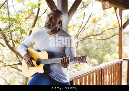 Calvo afroamericano anziano che suona la chitarra mentre si siede sulla ringhiera nel balcone alla cabina di ceppo Foto Stock