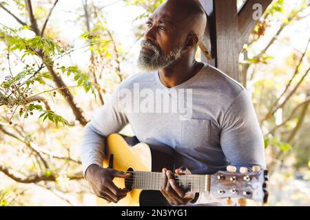 Uomo afroamericano calvo anziano che suona la chitarra mentre si siede dalla colonna nel balcone alla cabina del ceppo Foto Stock