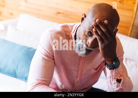 Primo piano di un anziano afro-americano calvo stressato con testa in mano seduta sul divano in una cabina di tronchi Foto Stock