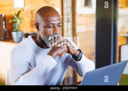 Serio uomo anziano afro-americano calvo con le mani aggrappate guardando il laptop in cabina di ceppo Foto Stock