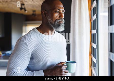 Uomo anziano afro-americano con una tazza di caffè che guarda attraverso la finestra alla cabina di tronchi Foto Stock