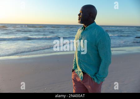 Uomo anziano afro-americano con le mani in tasca che guarda il mare contro il cielo al crepuscolo. Copia spazio, spiaggia, pensione, scenico, inalterato Foto Stock