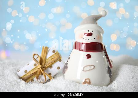 Pupazzo di neve decorativo vicino alla scatola regalo sulla neve artificiale contro le luci festive sfocate Foto Stock