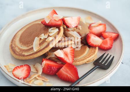 Frittelle vegane con fragole, burro di noci, patatine di cocco e sciroppo, close-uo. Foto Stock