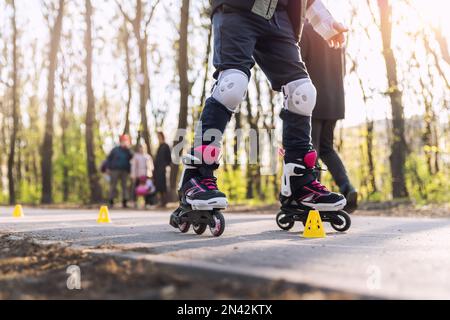 Un gruppo di bambini si diverte imparando la slalom dello skate a rotelle in linea con coni di plastica su strada nel parco cittadino all'aperto nella soleggiata giornata primaverile Foto Stock