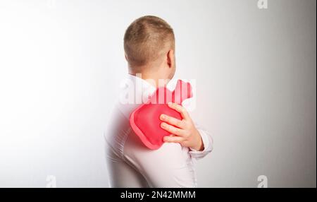 Un uomo tiene un tampone di riscaldamento medico rosso con acqua calda vicino alla sua articolazione della spalla. Spalline, artrite e tendinosi. Foto Stock