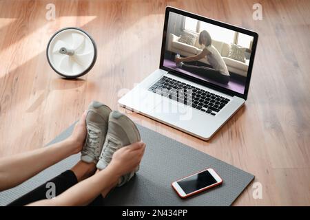 Flessibilità e resistenza. Adatta la giovane donna che stretching i muscoli a casa, facendo esercizi mentre guarda tutorial yoga online sul notebook. Concetto di fitness e home yoga Foto Stock