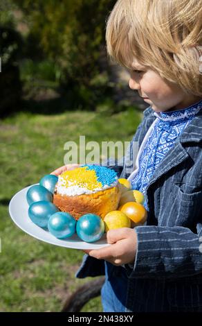 Nella soleggiata giornata primaverile, il bambino tiene un piatto con uova di Pasqua blu e gialle intorno ad una torta di Pasqua a colori della bandiera Ucraina. buona vacanza di pasqua, ukr Foto Stock