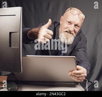 Lavorare in ufficio, un uomo anziano allegro con una barba grigia davanti a un notebook è soddisfatto del risultato. Foto Stock