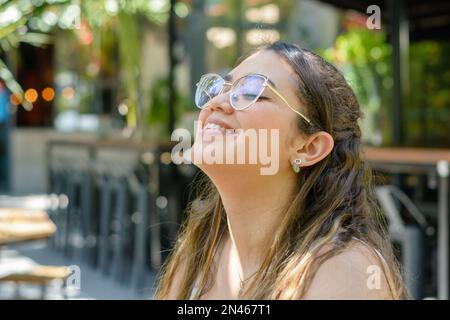 giovane donna venezuelana latina, con gli occhi chiusi, ridendo e respirando aria fresca a non stress mentre si aspetta il cibo seduto in un ristorante Foto Stock