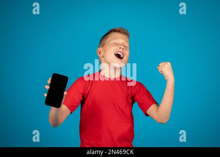 Ragazzo pieno di gioia che tiene uno smartphone vuoto e celebra il successo su sfondo blu Foto Stock