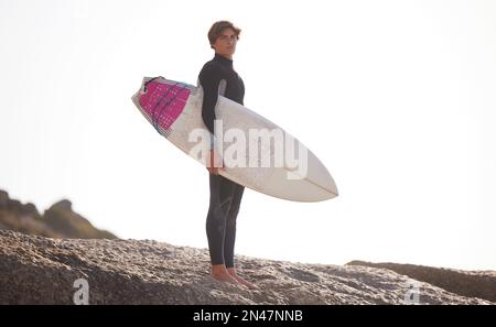 Surf, uomo con tavola da surf per sport all'aperto, rocce da spiaggia e viaggi con spazio mockup e natura. Avventura, sport estremi e ritratto surfista Foto Stock