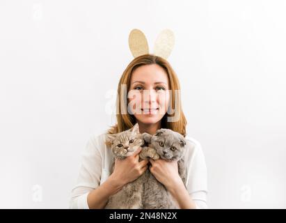 Giovani donne belle e positive che indossano orecchie conigliate e che si divertono con due gatti. Look minimalista sullo sfondo bianco. Concetto di Pasqua e delle feste Foto Stock