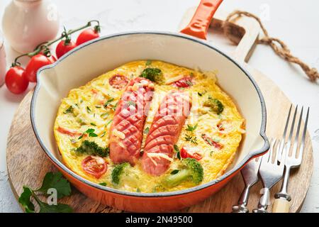 Omelette o frittata con due salsicce, formaggio fuso, pomodori ciliegini, broccoli e prezzemolo su padella rossa o teglia di ferro su fondo bianco di cemento vecchio Foto Stock