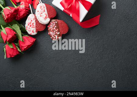 Fiori rossi di rosa, biscotti brownie e caramelle al cioccolato a forma di cuore, confezione regalo con vino su sfondo nero. Giorno di San Valentino, madri o donne g Foto Stock