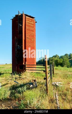 Vecchio silo rosso accanto a una recinzione in una fattoria in un campo di erba verde Foto Stock