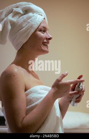 Foto verticale donna sorridente applicando crema anti-acne in faccia in bagno domestico, routine di cura della pelle Foto Stock