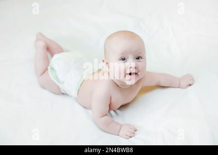 Il bambino giace sul pancino del letto con le gambe sollevate. Stendilo fuori sul vostro stomaco dopo il sonno. Un bambino in un pannolino su un letto bianco. Foto Stock