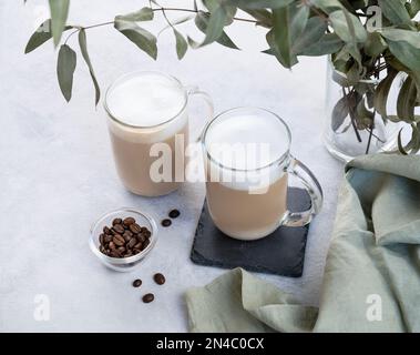 Due cappuccino o latte con schiuma di latte in bicchieri su sfondo chiaro con chicchi di caffè e ramificazione di eucalipto. Il concetto di colazione primaverile. Foto Stock