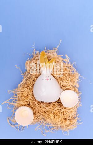 Un coniglietto pasquale bianco tratteggiato e gusci di uovo rotti in un nido su uno sfondo blu. Pasqua minima composizione creativa. Buona vacanza di Pasqua. Una greetina Foto Stock