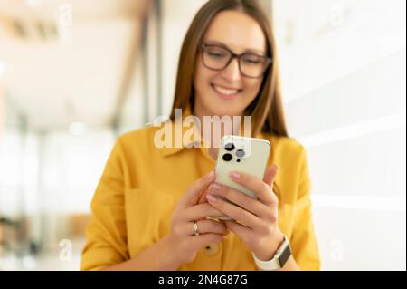 Focalizzazione selettiva su smartphone alla moda nelle mani di una donna d'affari in occhiali, dipendente di un ufficio femminile che utilizza il telefono per chattare online, messaggistica. Ragazza freelance che utilizza il telefono per lavorare in remoto Foto Stock